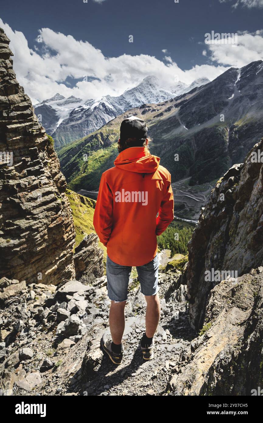 Porträt eines männlichen Wanderers in Shorts und einer Kappe von hinten steht auf hohen Felsen am Rand einer Klippe vor dem Hintergrund eines grünen Tals W Stockfoto