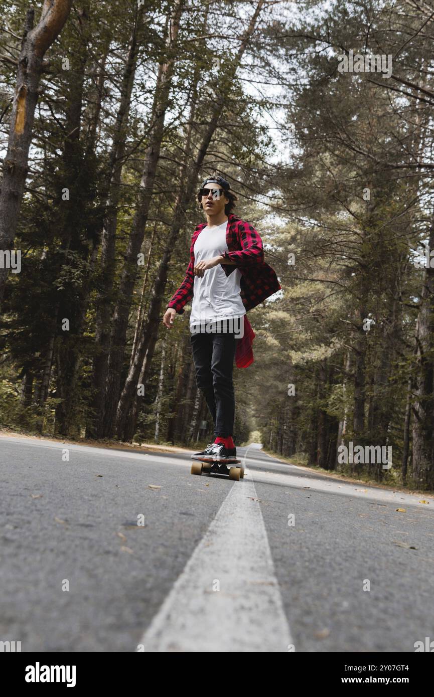 Ein junger Hipster in einer Mütze mit Sonnenbrille und einem karierten Hemd fährt auf seinem Skateboard auf einer Landstraße im Herbstwald. Das Konzept des Reisens Stockfoto