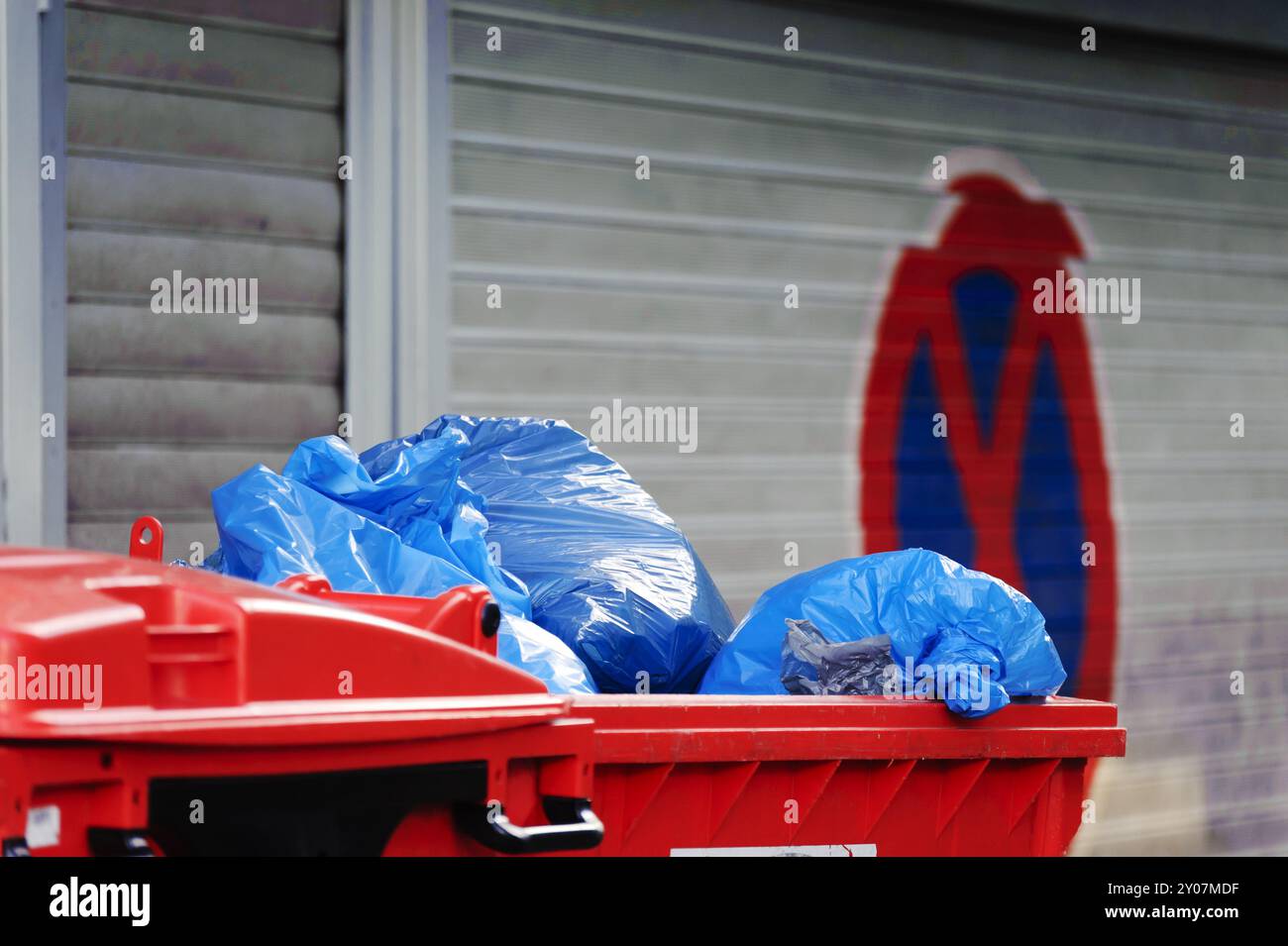 Rote Mülltonne mit blauen Plastiktüten vor einer Rollladentür Stockfoto