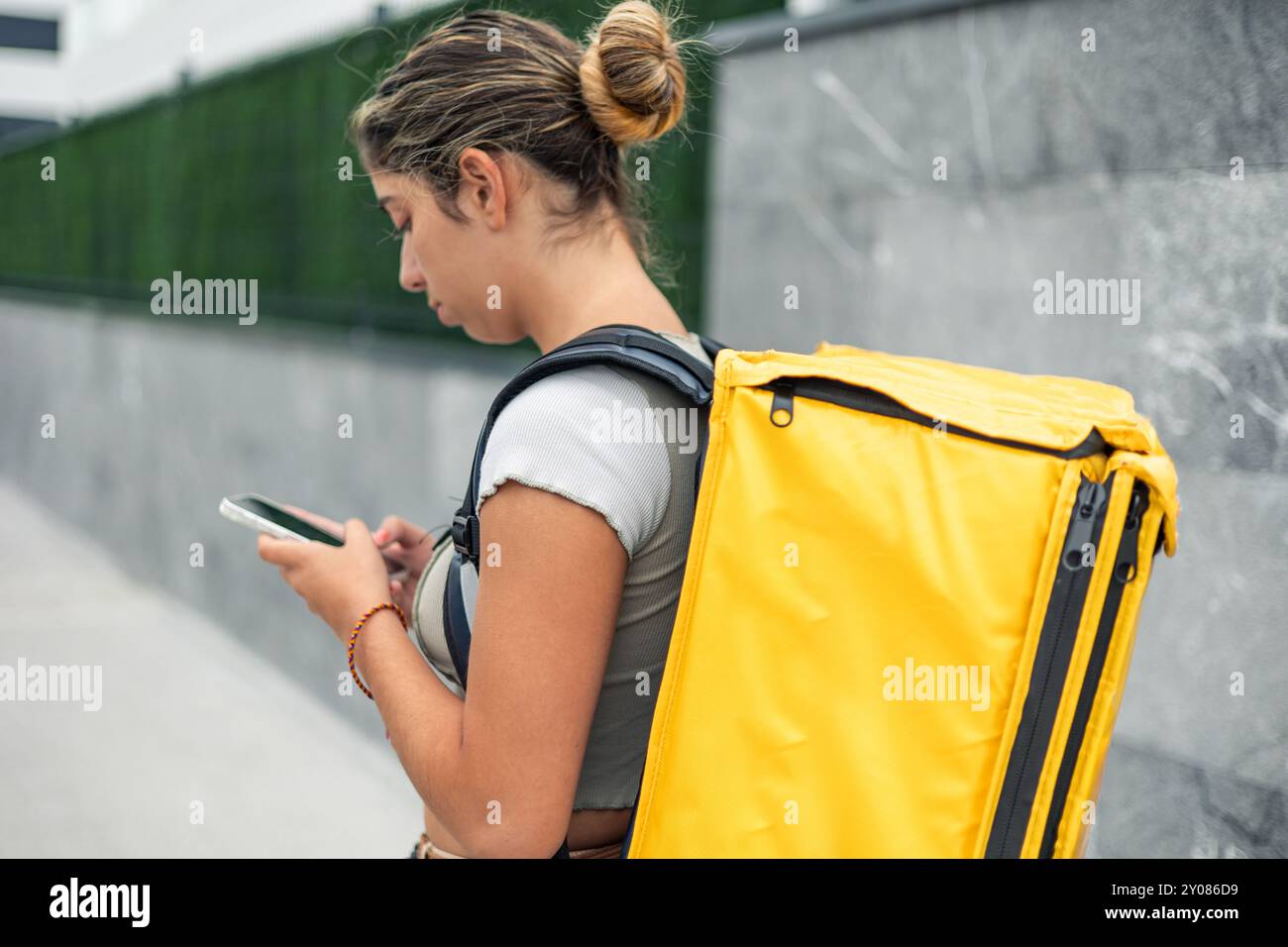 Die hispanische Essenszustellerin mit gelbem Rucksack fragt ihr Smartphone, um die Adresse des Kunden zu überprüfen, an den sie die Bestellung liefern muss. Stockfoto