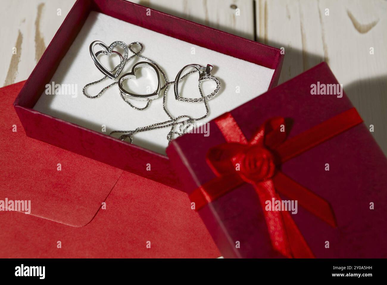 Großaufnahme von silbernen Herzanhängern in einer roten Geschenkbox und einem roten Umschlag auf einem hölzernen Hintergrund Stockfoto