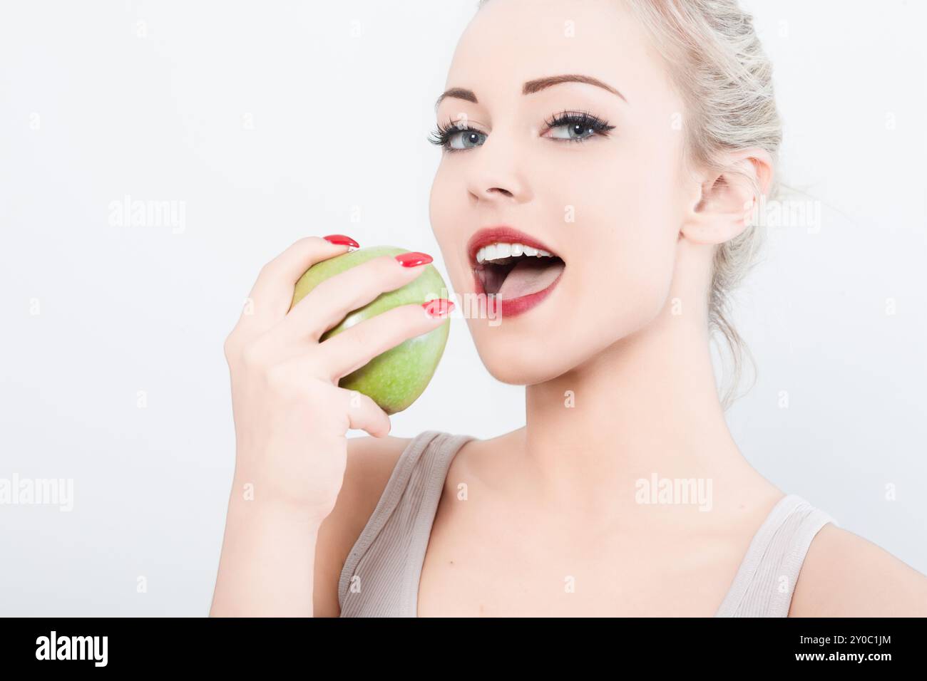 Blonde Frau mit rotem Lippenstift hält einen grünen Apfel, bereit zum Beißen. Ihr fröhlicher Ausdruck zeigt Freude am gesunden Essen. Das Bild zeigt den Wunsch nach Nutriti Stockfoto