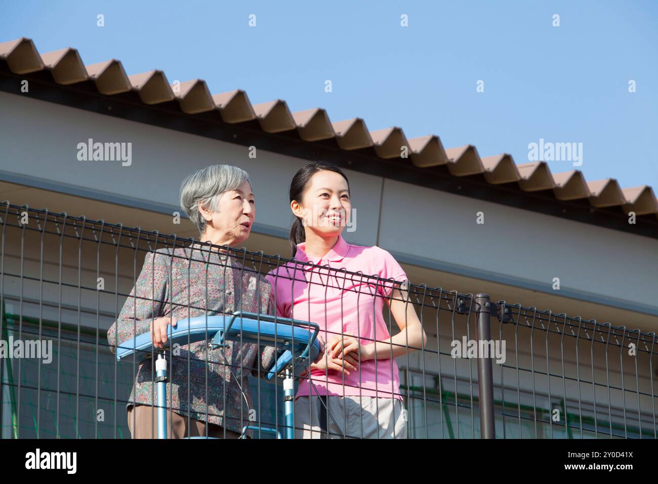 Eine ältere Frau, die von einem Balkon aus den Blick nach draußen genießt, begleitet von ihrer Betreuungsperson Stockfoto