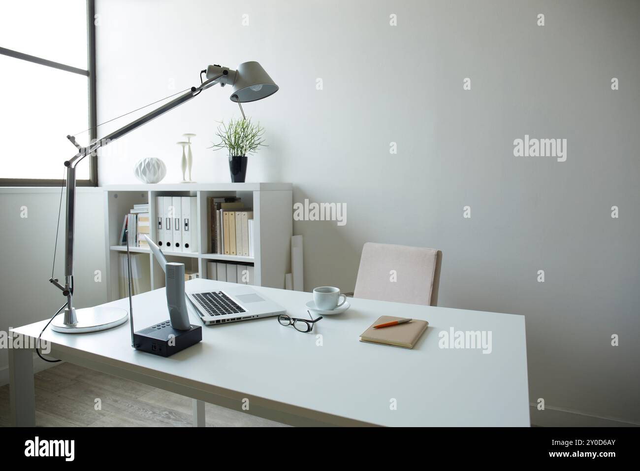 Ein Schreibtisch mit weißen Möbeln und Dokumenten, eine Tasse Kaffee, Gläser und ein Laptop auf dem Bücherregal in einem weißen Raum Stockfoto