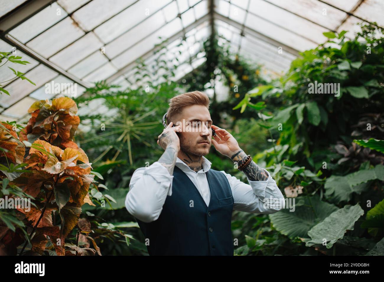 Gutaussehender Mann mit Tätowierung, der Meditationsmusik im botanischen Garten zuhört, inmitten von üppig grünem Laub. Er kümmert sich um seine psychische Gesundheit Stockfoto