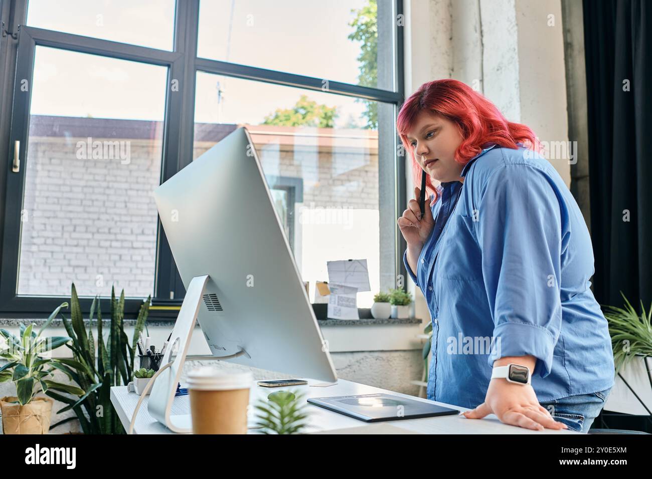 Eine große Frau in einem blauen Hemd betrachtet ihre Arbeit vor einem Computermonitor. Stockfoto