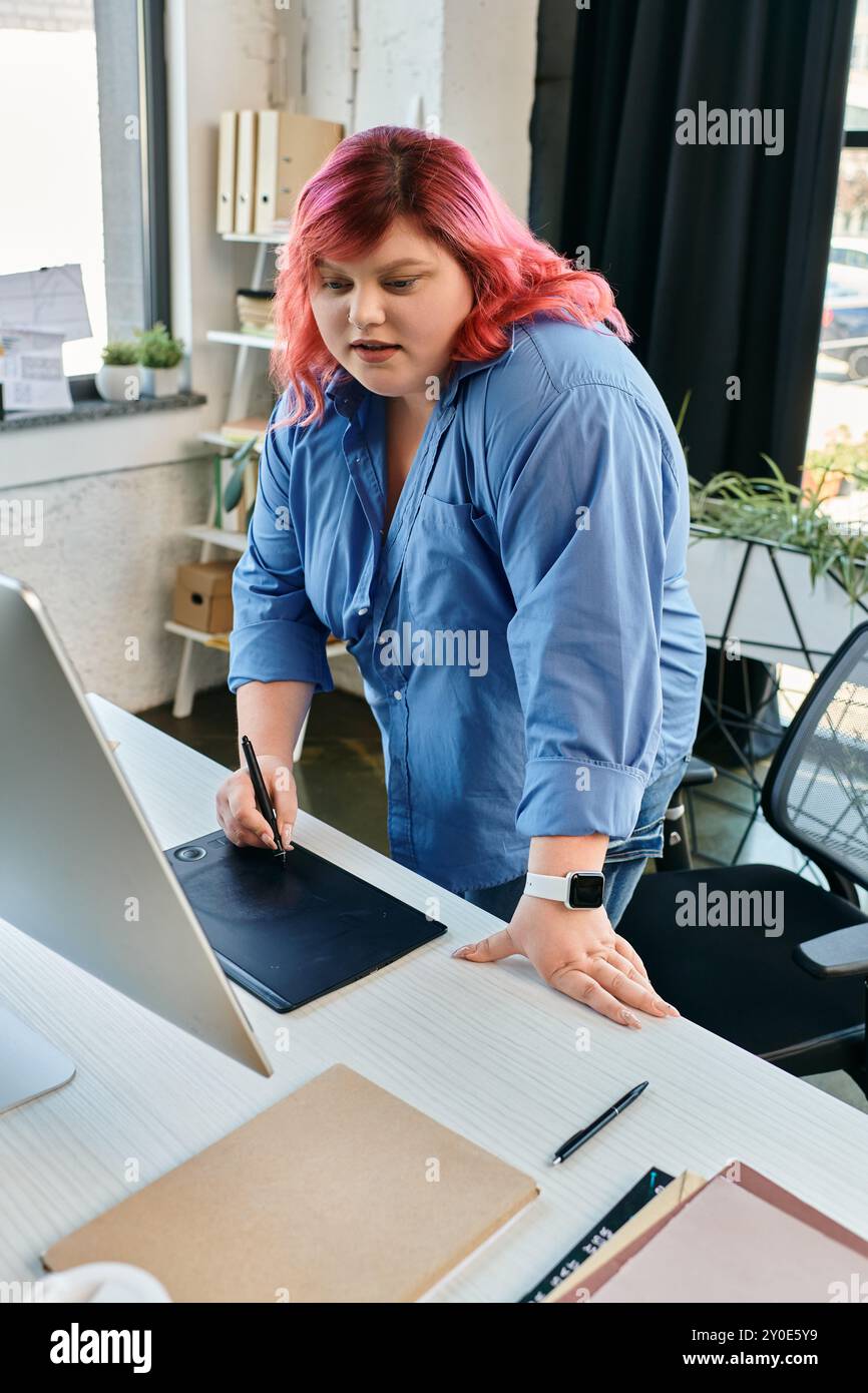 Eine Frau in Übergröße mit hellrosa Haaren und blauem Hemd verwendet einen Stift auf einem digitalen Zeichentablett. Stockfoto