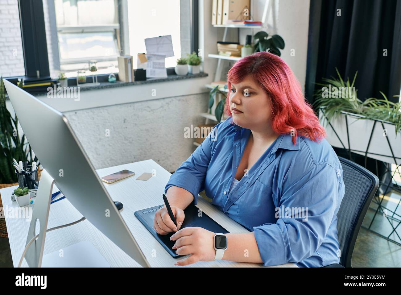 Eine Frau in Übergröße mit leuchtendem rosa Haar arbeitet mit einem Stift und Tablet an einem digitalen Design. Stockfoto