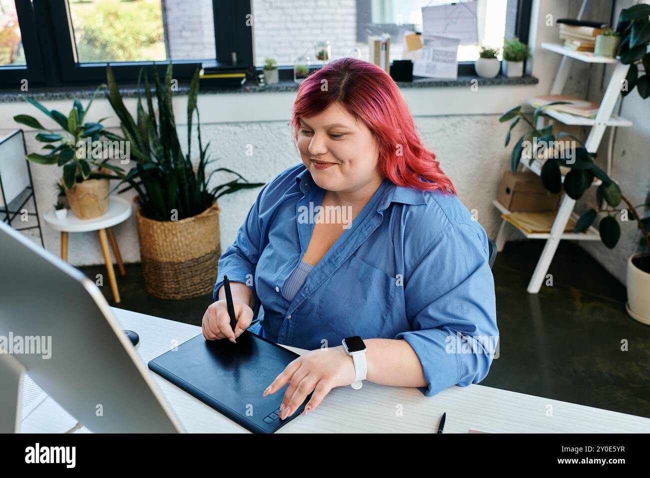 Eine große Frau mit knallroten Haaren zeichnet auf einem digitalen Tablet, während sie an ihrem Schreibtisch arbeitet. Stockfoto