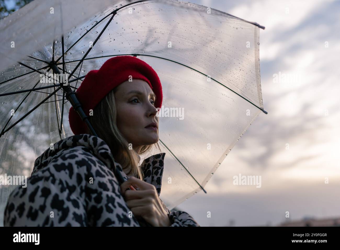 Eine Frau, die einen roten Hut und einen Mantel mit Leopardenmuster trägt, hält einen klaren Regenschirm. Der Schirm ist offen und die Frau blickt in den Himmel. Die Stockfoto