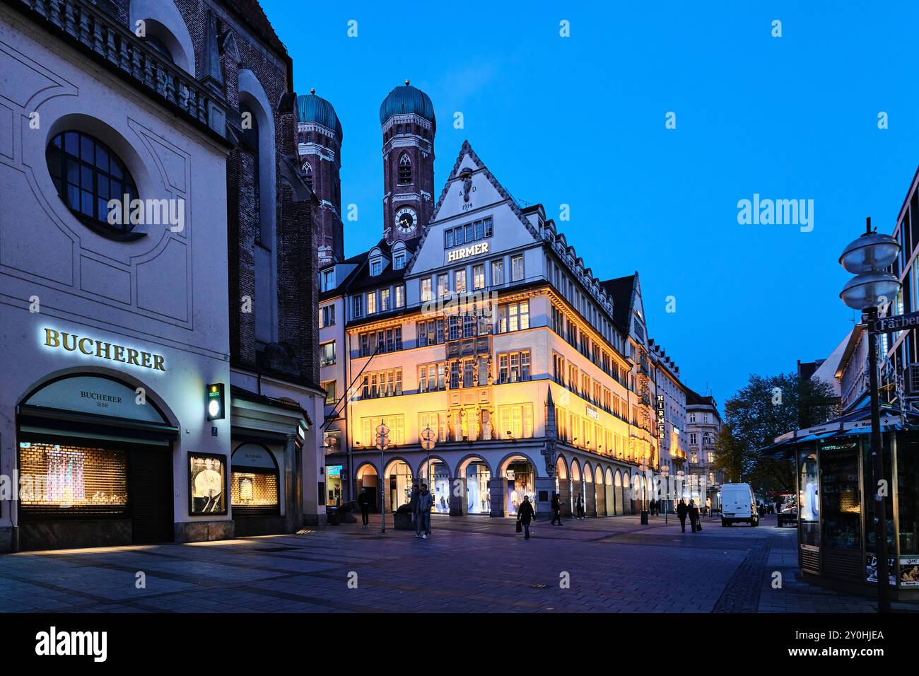 München - 18. April 2024: Außenansicht des größten Herrenmode-Hauses Hirmer. Die Türme der Frauenkirche Stockfoto