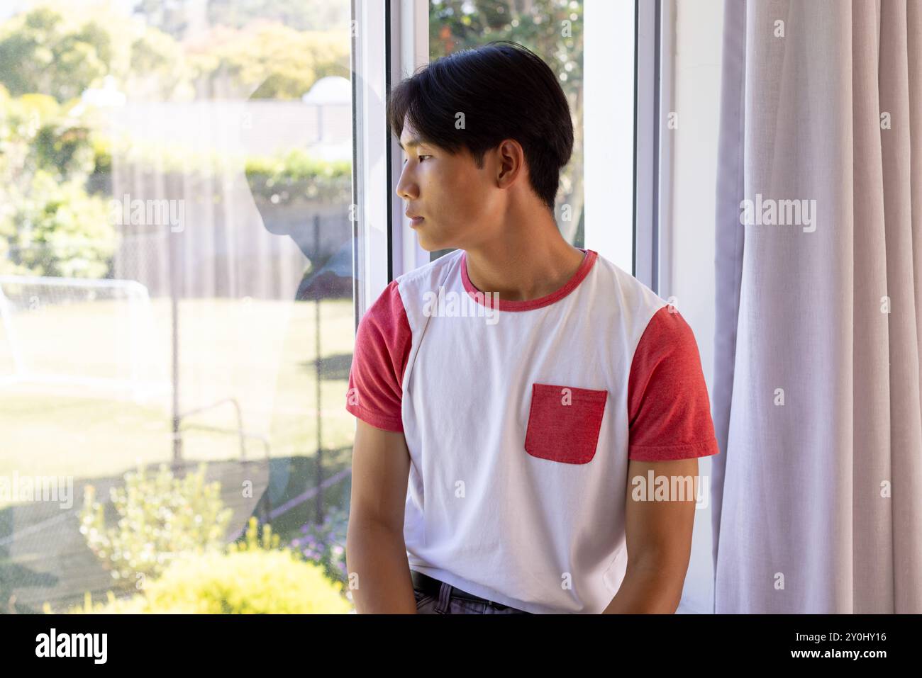 Aus dem Fenster schaut ein junger asiatischer Mann in lässigem Hemd nach draußen Stockfoto