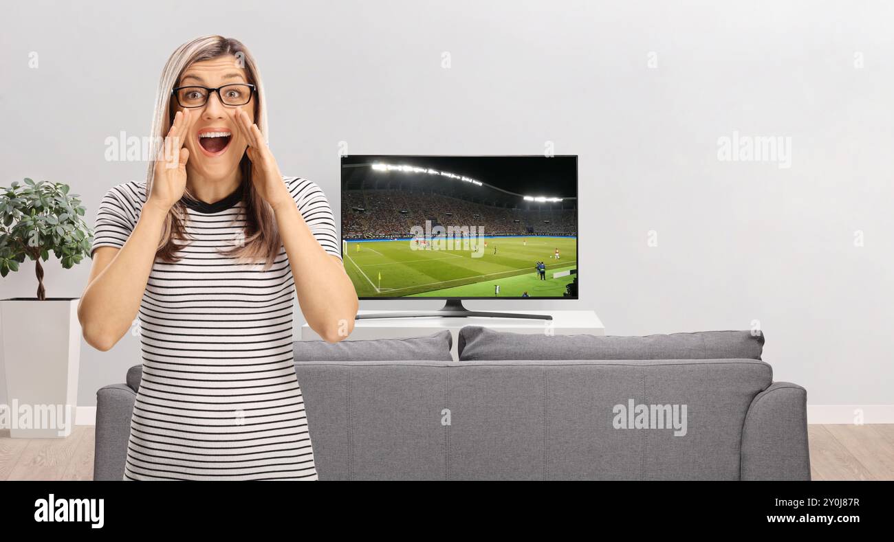 Blonde Frau mit Brille, die Fußball schaut und vor dem fernseher zu Hause brüllt Stockfoto