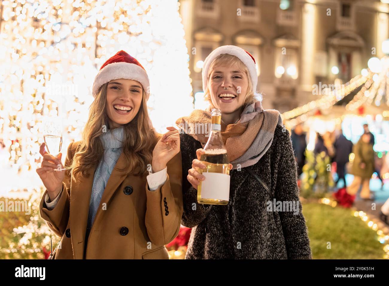 Zwei fröhliche junge Frauen in Weihnachtsmützen genießen festliche Drinks auf einem Weihnachtsmarkt. Umgeben von Weihnachtslichtern und Dekorationen - Freude und Wärme von Stockfoto