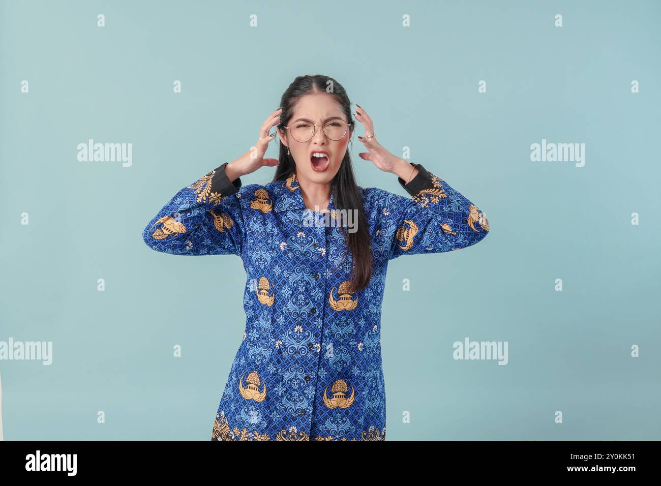 Frustrierte indonesische Regierungsangestellte in blauer Batikuniform, wütend und hebt ihre Hände in die Luft. PNS-Beamtenkonzept. Stockfoto