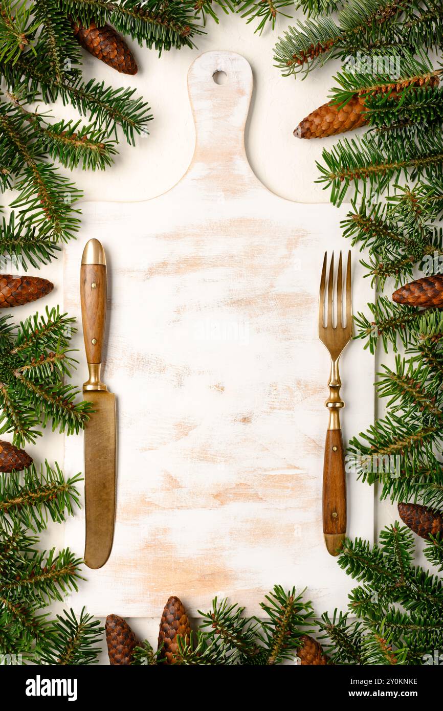 Hintergrund des Weihnachtsmenüs mit weißem Holzschneidebrett, Messer, Gabel und frischen Tannenzweigen mit Kegeln. Weihnachten oder Neujahr Hintergrund. Kreatives Mockup für Winterferien Stockfoto
