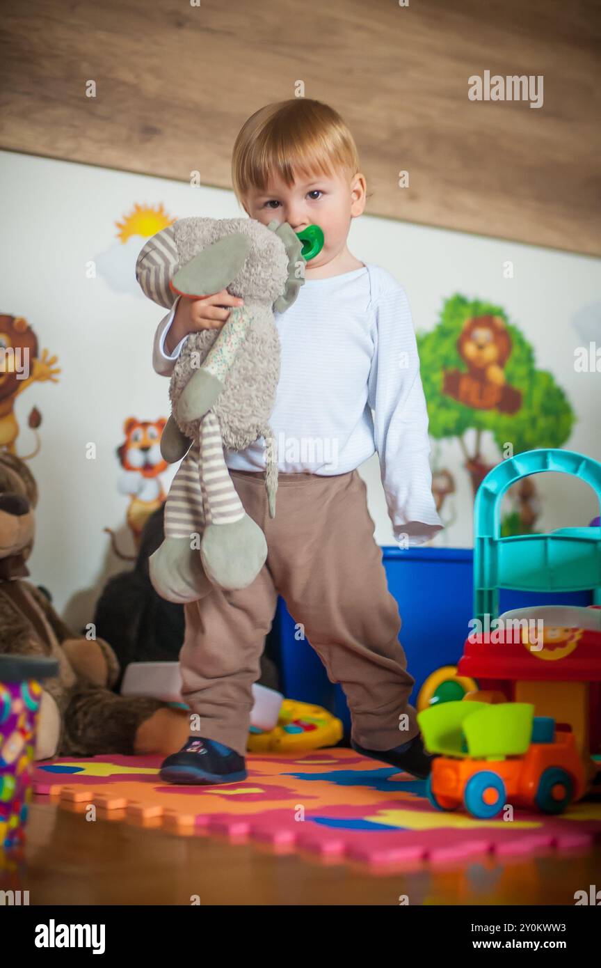 In einem lebendigen Spielzimmer voller Spielzeuge steht ein fröhlicher Junge stolz in der Hand seines geliebten Stofftieres. Sanfte Farben und verspielte Designs umgeben Stockfoto