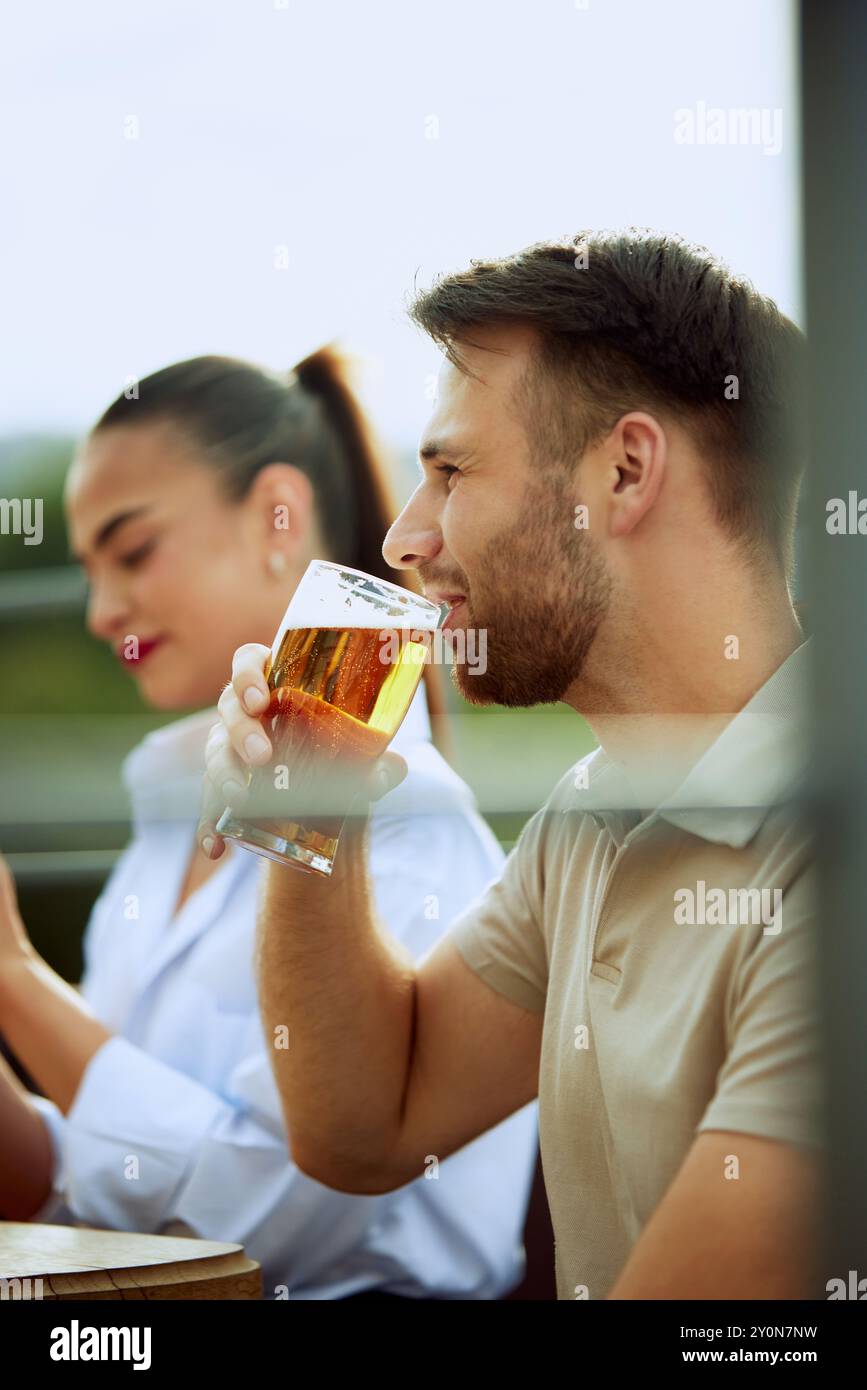 Junger bärtiger Mann genießt kaltes Bier in entspannter Atmosphäre im Freien und genießt Momente. Frau sitzt in der Nähe. Festliche Atmosphäre. Stockfoto