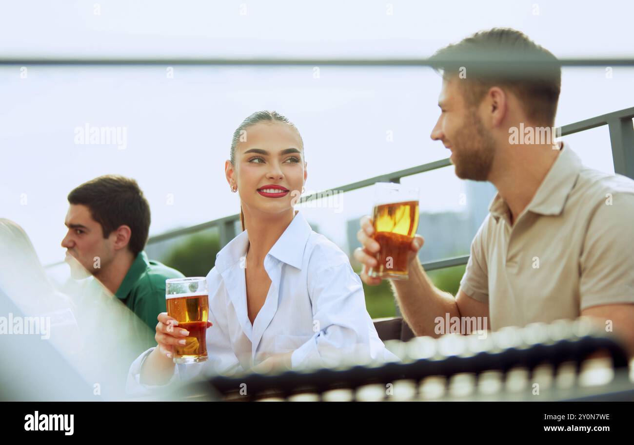 Junger bärtiger Mann genießt kaltes Bier in entspannter Atmosphäre im Freien und genießt Momente. Frau sitzt in der Nähe. Festliche Atmosphäre. Stockfoto