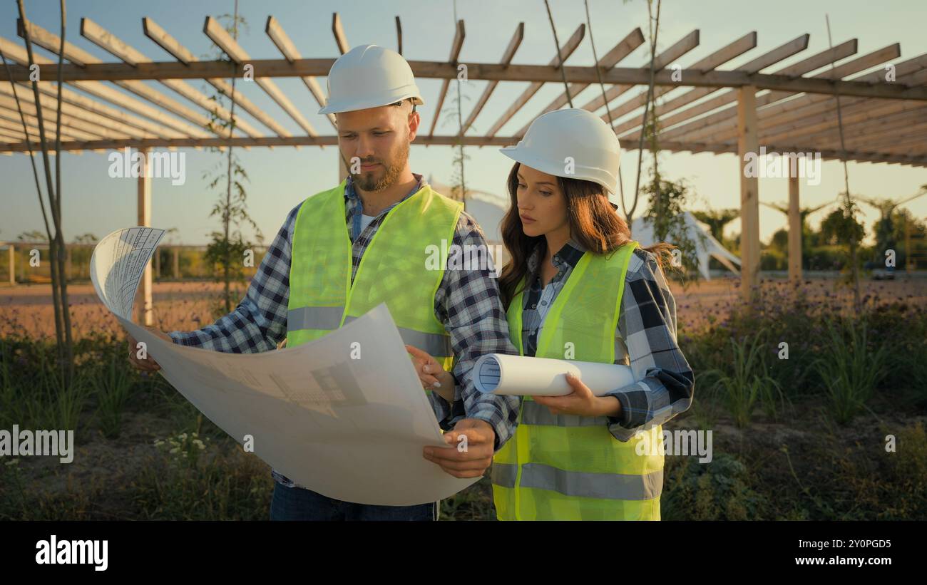 Zwei kaukasische Arbeiter Uniform Hardhat weibliches Mädchen männlicher Mann Mann Ingenieure Partner halten Blaupausen ökologischem Erbauer zeigt Baustellenspezialisten Stockfoto