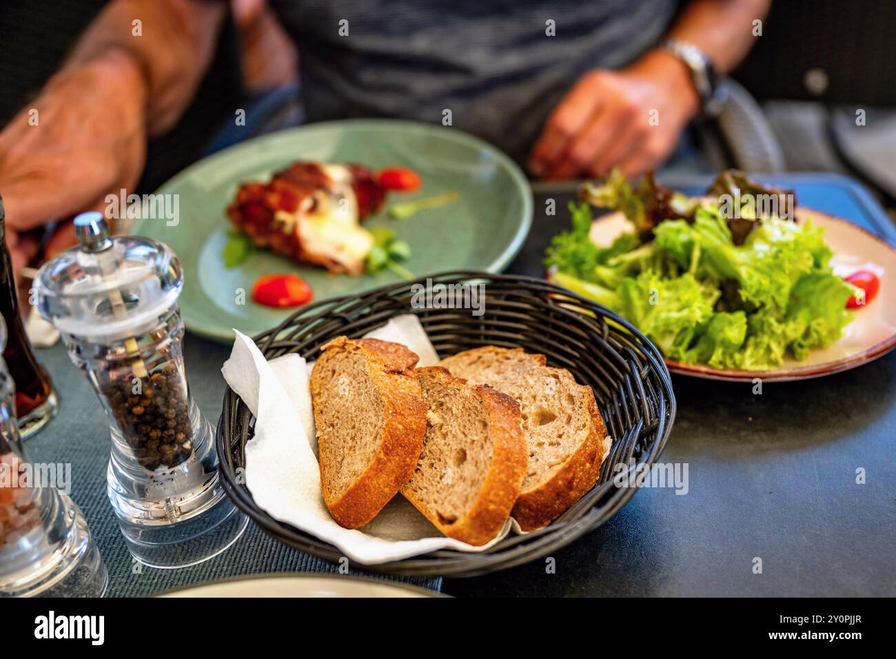 Der Mann isst im Restaurant, auf dem Tisch ist Brot mit Serviette im Korb, Salat, Shaker und ein Stück Fleisch auf dem dritten Teller. Stockfoto