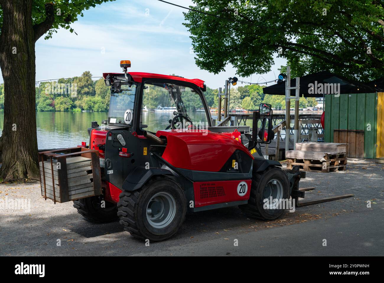 Ein kompakter roter Gabelstapler parkt am Wasser, umgeben von Grün und städtischen Strukturen an einem klaren, sonnigen Tag. Stockfoto