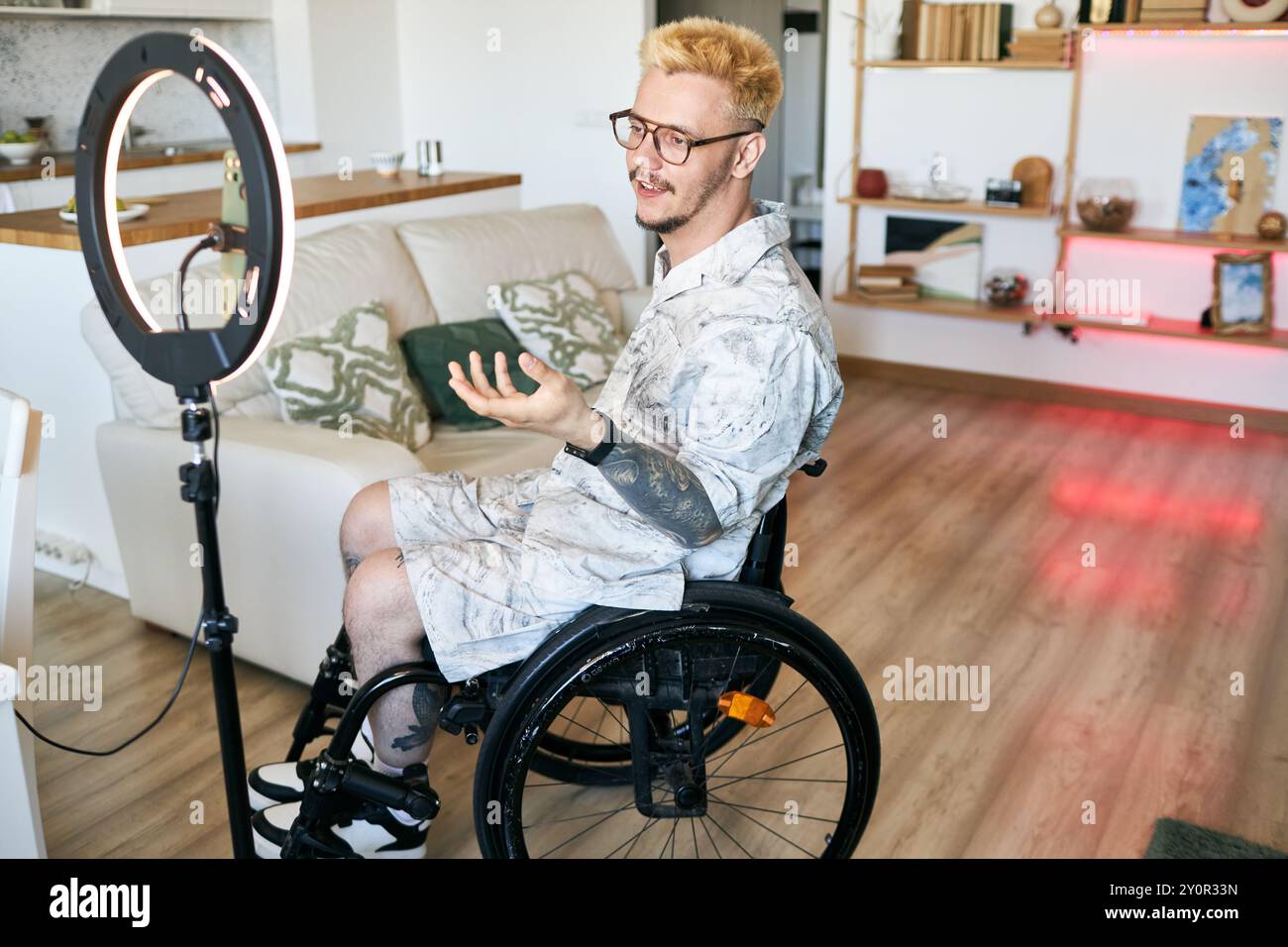 Mann im Rollstuhl nimmt Lehrvideos in moderner Wohnzimmerumgebung mit Ringlicht und Kamera auf, trägt eine Brille und spricht selbstbewusst Stockfoto