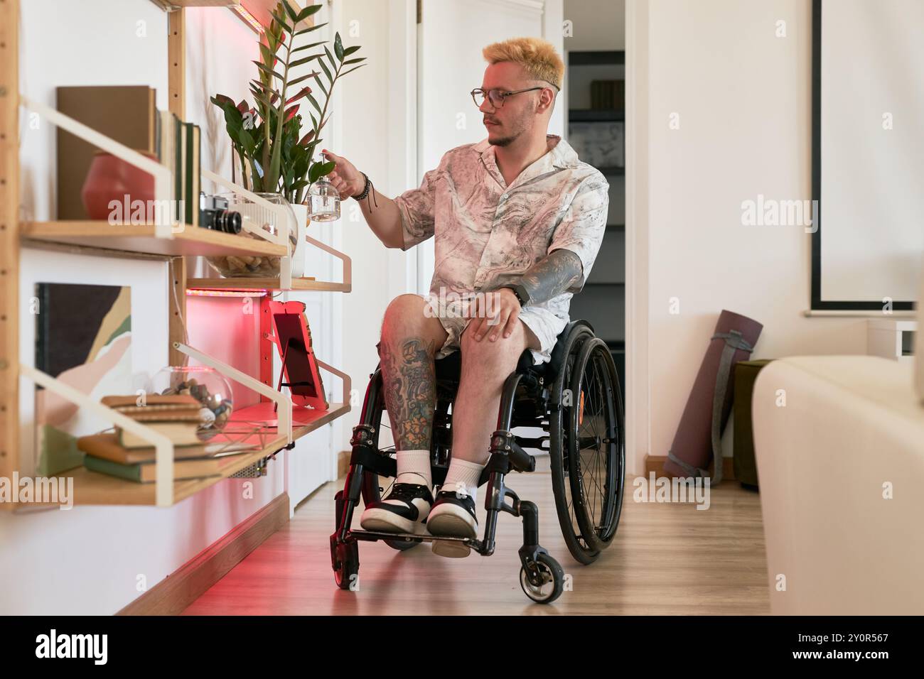 Kaukasischer Mann im Rollstuhl, der durch eine moderne Wohnung fährt und nach Artikeln im Regal sucht. Wohnzimmer mit Holzfußboden und stilvoller Einrichtung für eine einladende Atmosphäre Stockfoto