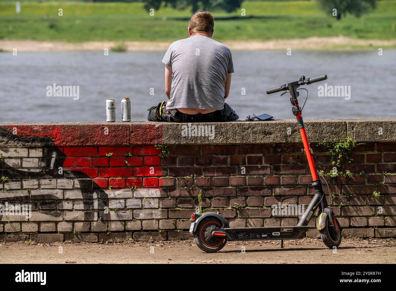 Mann, der an einer Mauer sitzt, am Rhein, trinkt Bier aus Dosen, kommt mit einem E-Roller an, Stockfoto