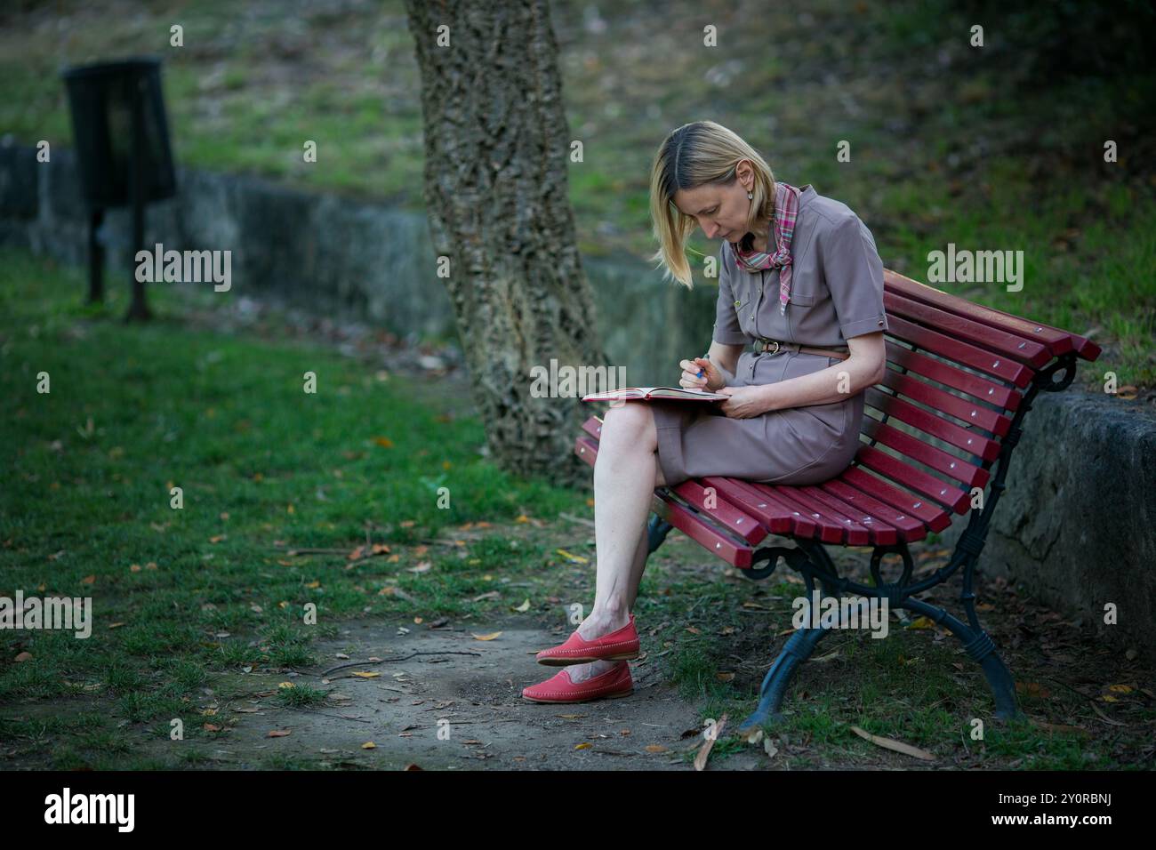 Eine Frau sitzt auf einer Parkbank und schreibt mit einem Stift in ein Notizbuch oder Tagebuch. Stockfoto