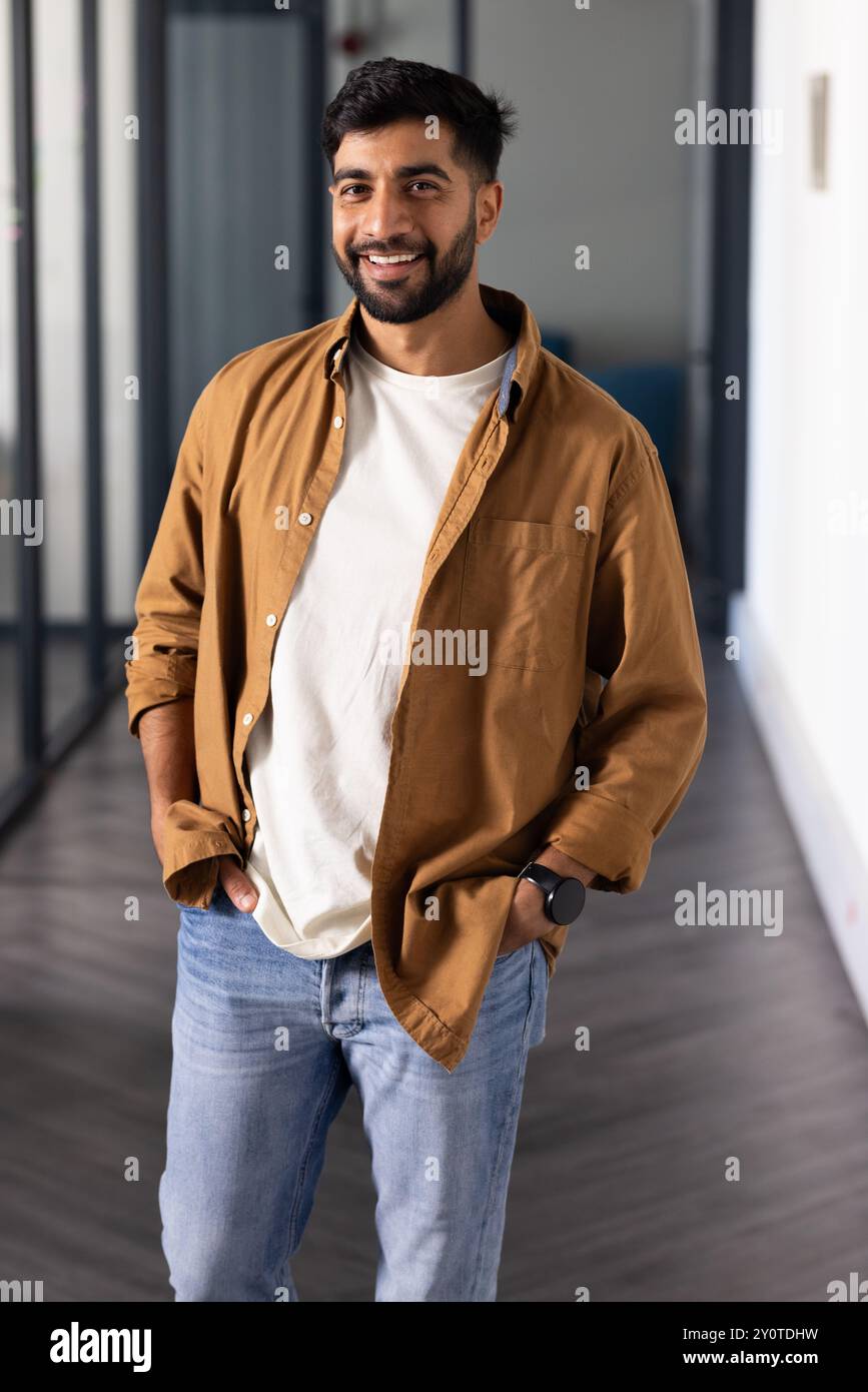 Ein lächelnder Indianer steht im Flur des Büros mit Händen in Taschen und trägt legere Outfits Stockfoto