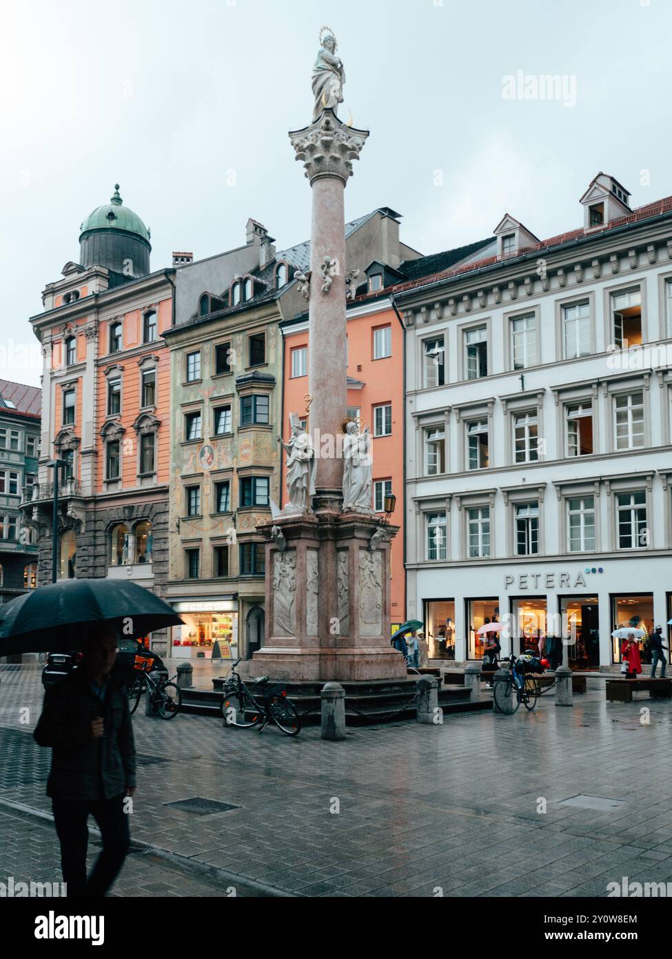 Eine Einzelfigur spaziert unter einem Sonnenschirm auf einem pulsierenden Stadtplatz mit dekorativer Säule und Skulpturen, umgeben von charmanter Architektur und Geschäften. Stockfoto