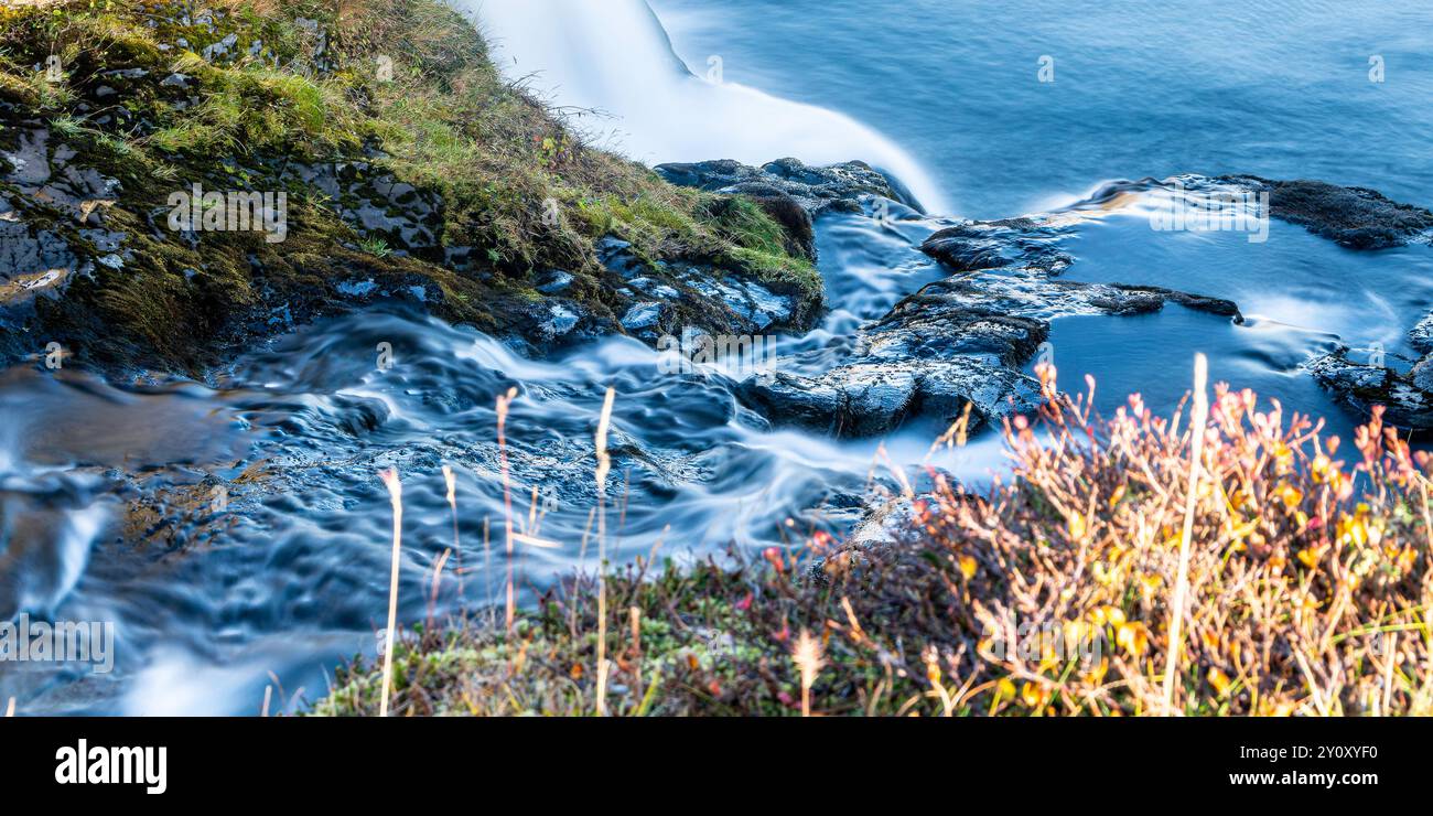 Ein Wasserstrom fließt über Felsen und Gras. Seideneffekt. Das Wasser ist ruhig und klar und die Felsen sind zerklüftet und rau. Das Gras ist hoch Stockfoto
