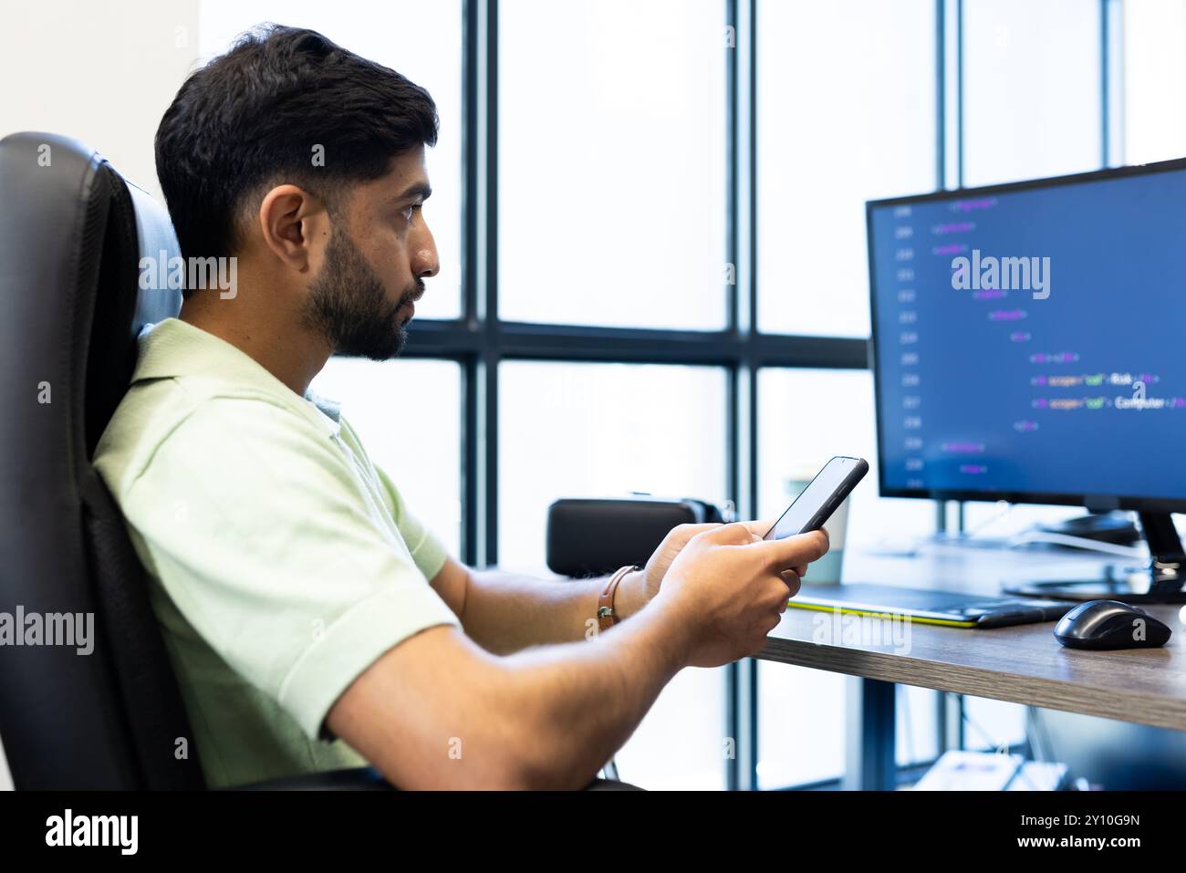 Mit Smartphone sitzt ein indischer Mann am Schreibtisch mit einem Computerbildschirm, der Code anzeigt Stockfoto