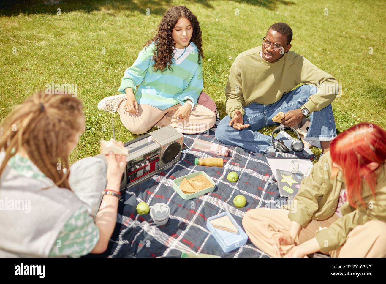 Verschiedene Gruppen von Studenten treffen sich zum Picknick auf dem grünen Rasen, unterhalten sich und genießen sich in Gesellschaft, während sie bei Sonnenschein im Freien Snacks essen Stockfoto