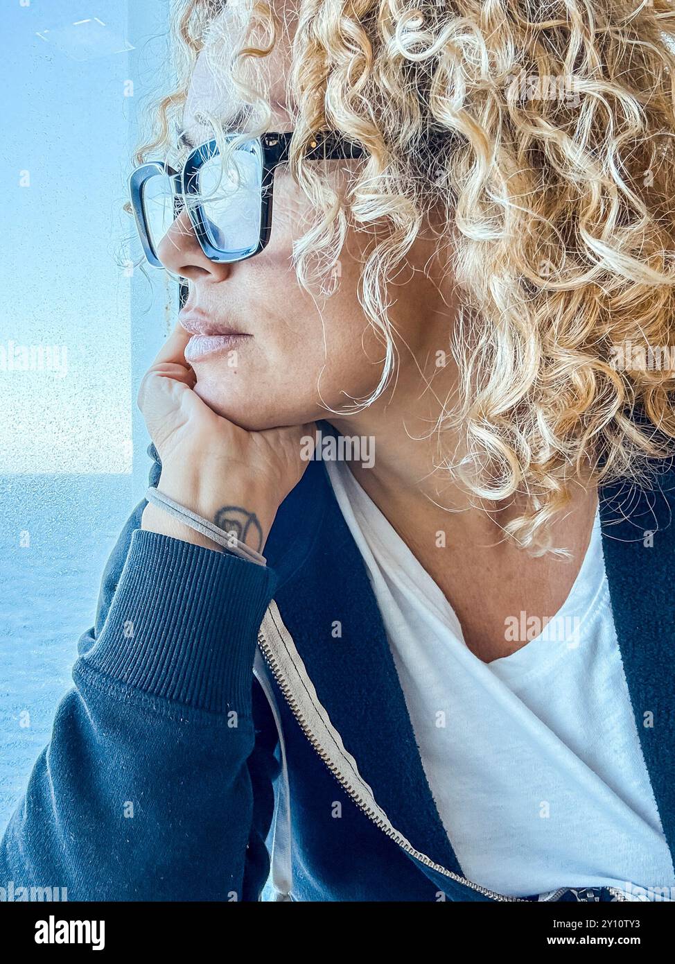 Schöne Frau mit Brille, die Freizeit mit Blick auf das Meer verbringt. Sie kümmert sich um sich, entspannt. Genießen Sie die Aussicht, schauen Sie durch das Fenster. Blondes und lockiges Weibchen mittleren Alters sieht nachdenklich aus. Ruhiger Morgen. Stockfoto