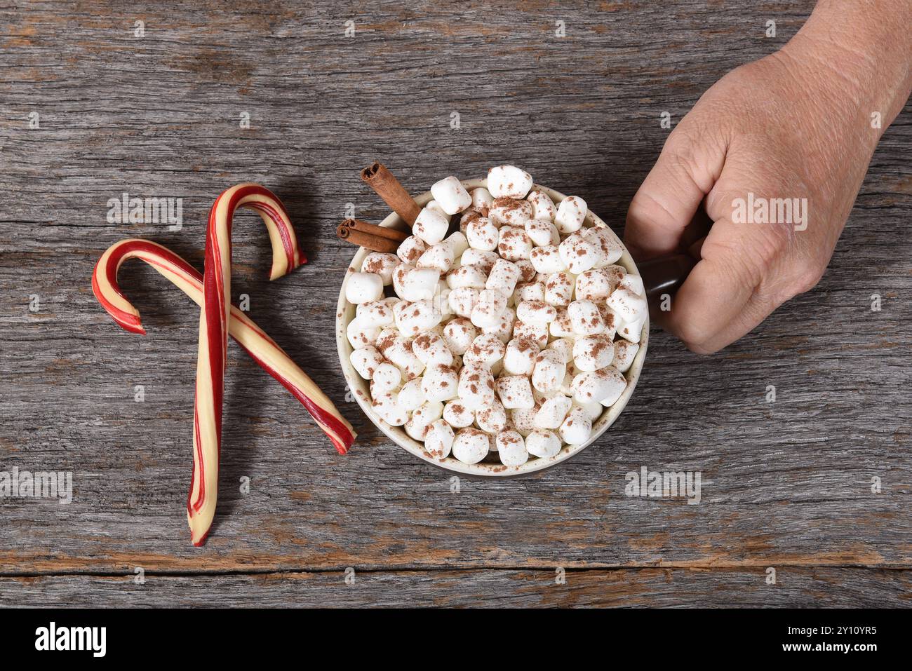 Blick von oben auf Santa Claus Hand mit einer großen Tasse heißen Kakao mit Marshmallows und Zimt. Zwei gekreuzte Süßigkeiten stehen neben dem Becher. Stockfoto