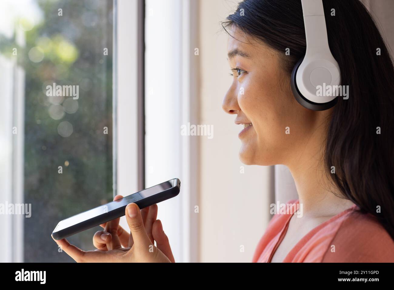 Sie trägt Kopfhörer, asiatische Frau verwendet Smartphone für Sprachbefehle in der Nähe des Fensters Stockfoto