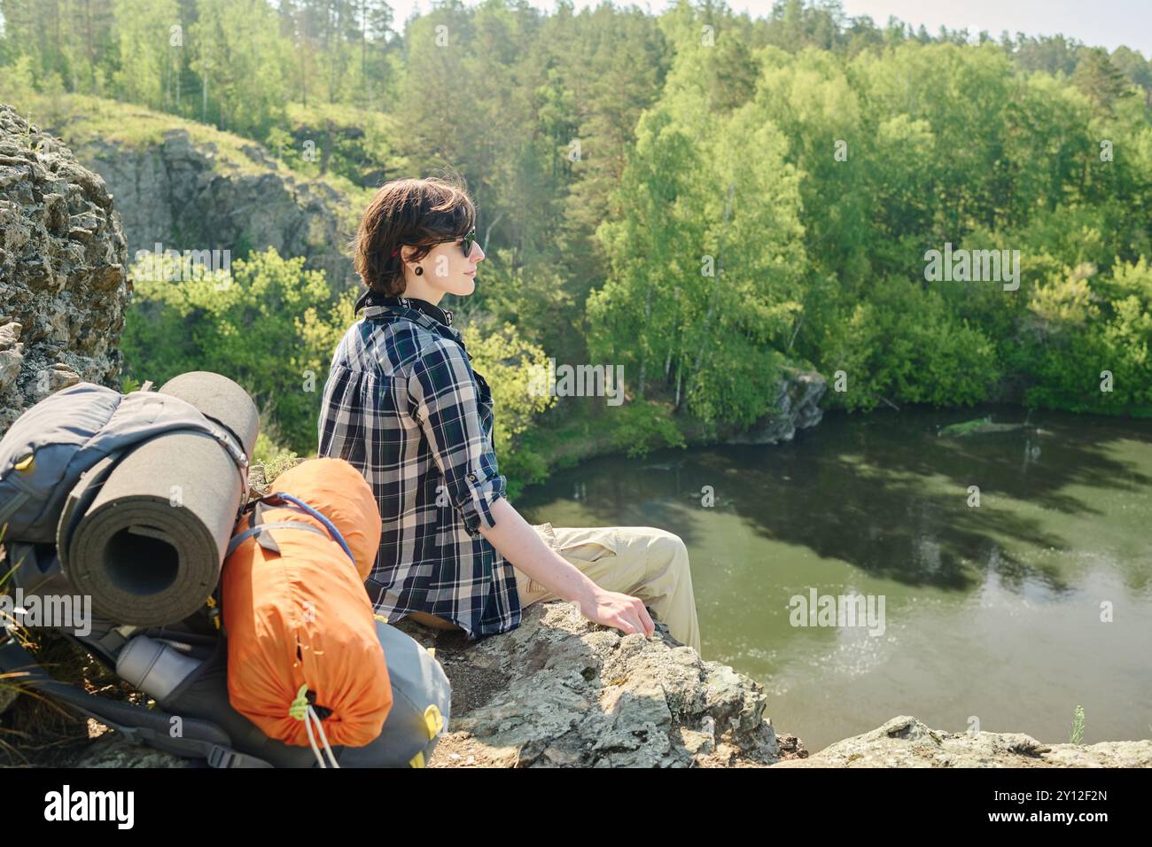 Junge, erholsame Wanderer in kariertem Hemd und Sonnenbrille, die auf dem Gipfel des Berges sitzen und während der Fahrt den Fluss bewundern Stockfoto