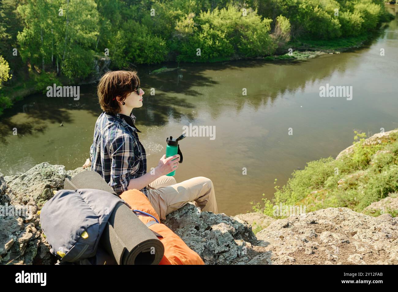 Junge Brünette weibliche Camper mit einer Flasche Wasser, die auf Klippen am Fluss sitzt und die Ruhe und die wunderbare Landschaft genießt Stockfoto