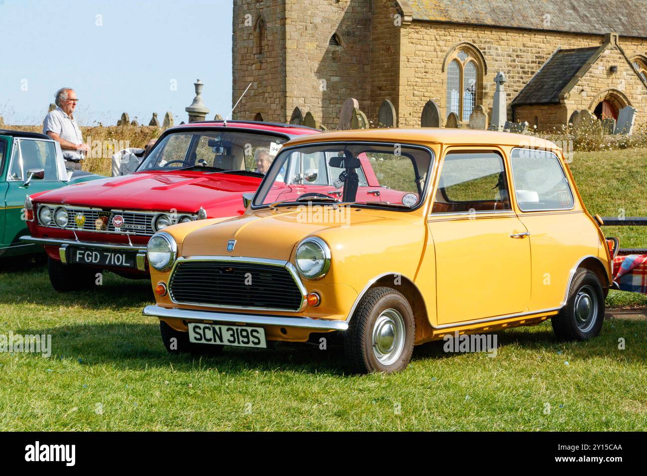 Klassischer gelber Vintage british leyland 1977 Mini 1000 und 1972 roter rover 2000 P6 geparkt auf Grasfeld und alten Steinkirchhintergrund. Stockfoto