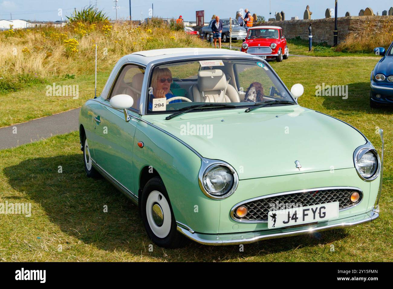 Ein klassischer hellgrüner Nissan Figaro, der an einem sonnigen Tag mit Menschen und einem anderen Oldtimer im Hintergrund gefahren wird. Stockfoto