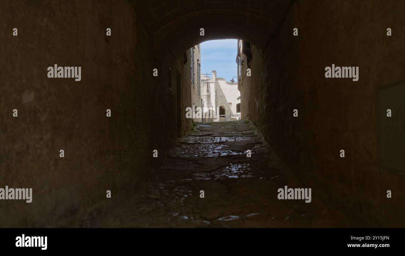 Eine enge, kopfsteingepflasterte Gasse in matera, basilicata, italien, bietet einen hellen Blick auf historische Steinhäuser unter einem klaren blauen Himmel durch einen Bogen Stockfoto