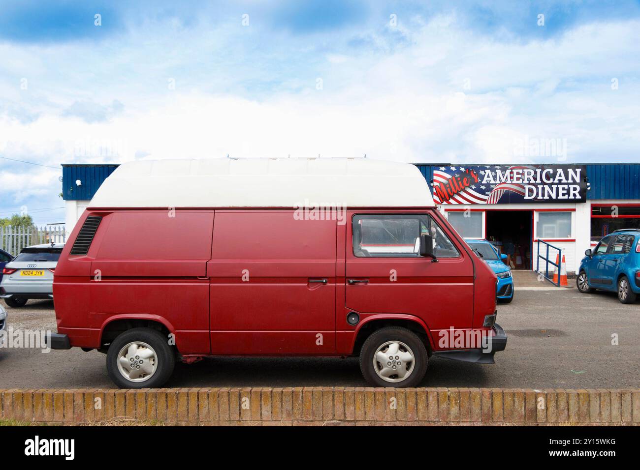 Roter Vintage Volkswagen Transporter T3 Hi Top Van, der vor einem amerikanischen Diner geparkt ist, mit blauem Himmel und mehreren Autos im Hintergrund. Stockfoto