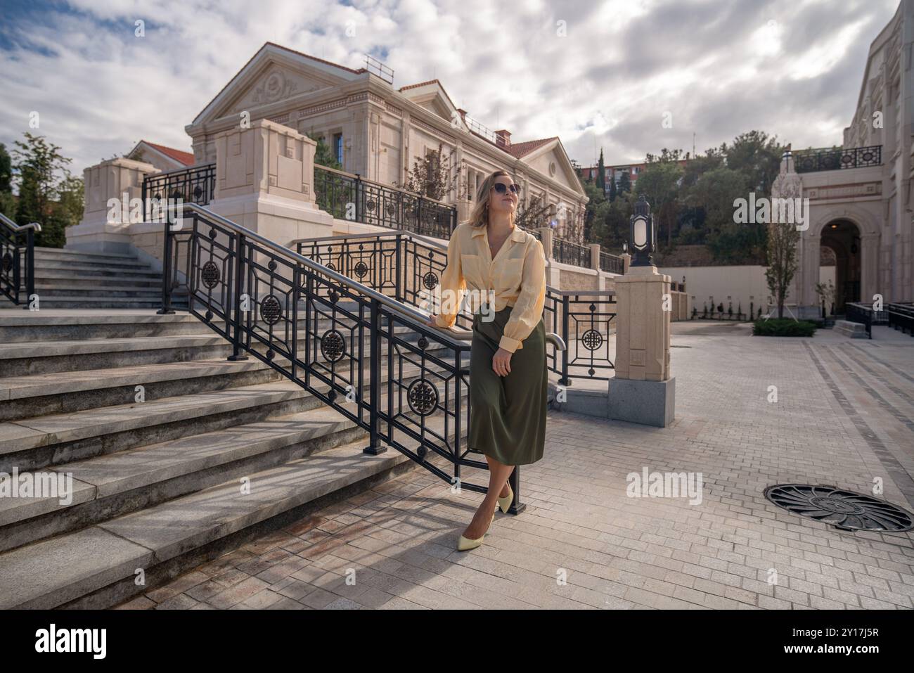 Eine Frau in beigem Hemd und grünem Rock steht auf der Treppe eines großen Steingebäudes. Sie lehnt sich auf das schwarze Geländer. Stockfoto