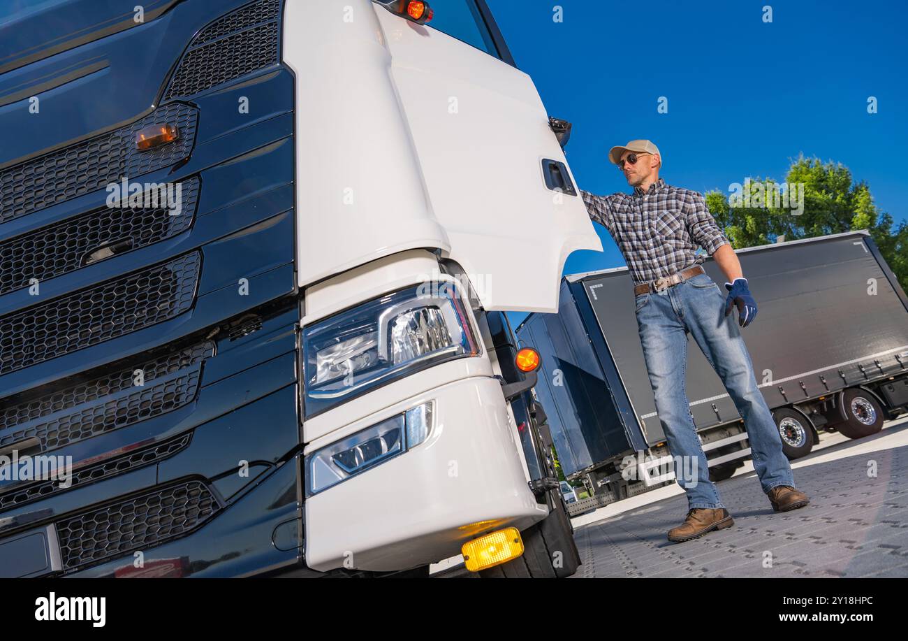 Ein Lkw-Fahrer, der ein kariertes Hemd und Handschuhe trägt, überprüft an klaren Tagen die Außenseite seines Lkws auf einem hellen, offenen Parkplatz. Stockfoto