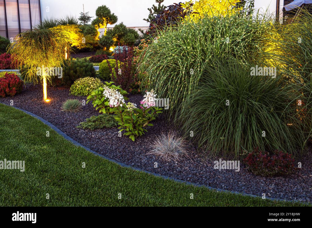 Ein wunderschön gestalteter Garten bietet üppiges Grün und farbenfrohe Blumen, beleuchtet von sanften Lichtern in der Abenddämmerung, was eine einladende Atmosphäre schafft. Stockfoto