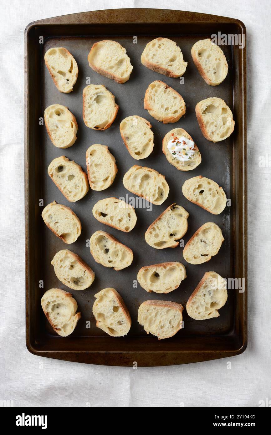 Ein Baguette, das in Scheiben geschnitten und auf einem Backblech verteilt wird, um das Knoblauchbrot zuzubereiten. Eine Scheibe enthält Gewürze und Kräuter und Topping bereits hinzugefügt. V Stockfoto