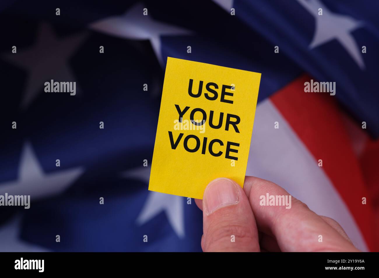 Eine Hand, die eine gelbe Papiernote mit dem Slogan „Use Your Voice“ vor der Flagge der USA hält. Stockfoto