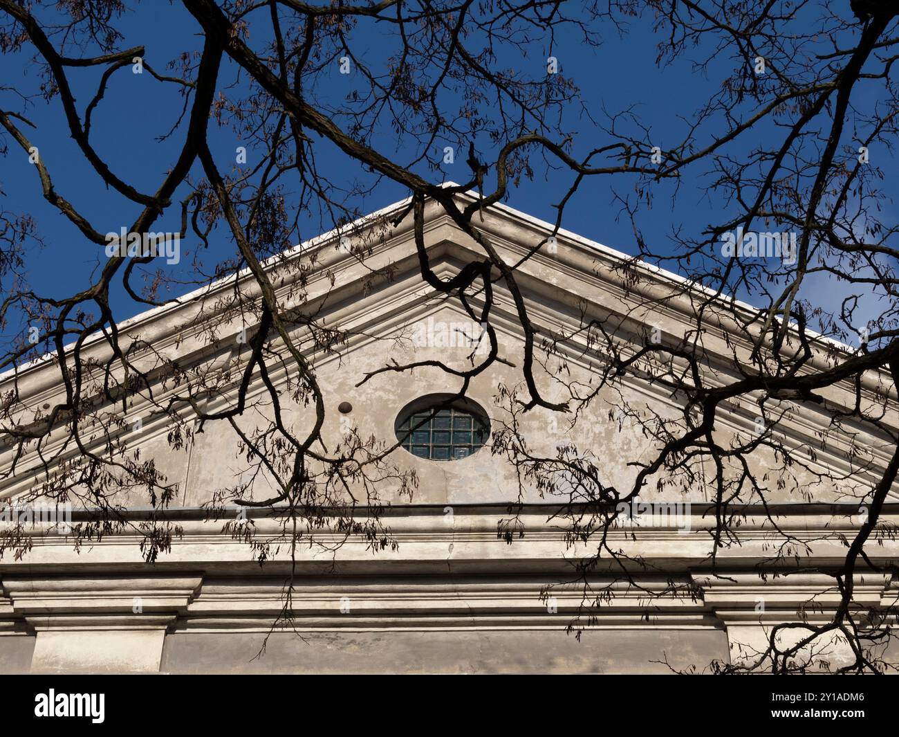 Oberer Teil einer alten Kapelle, verwitterte Farbe an der Wand, Zweige ohne Blätter im Vordergrund, blauer Himmel. Stockfoto